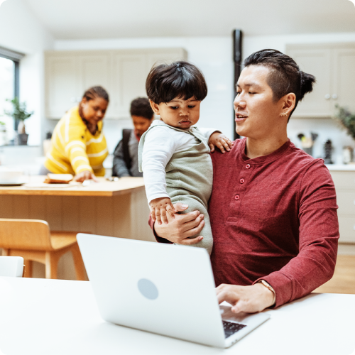 ’A proud father holds his infant son while sitting at a laptop’